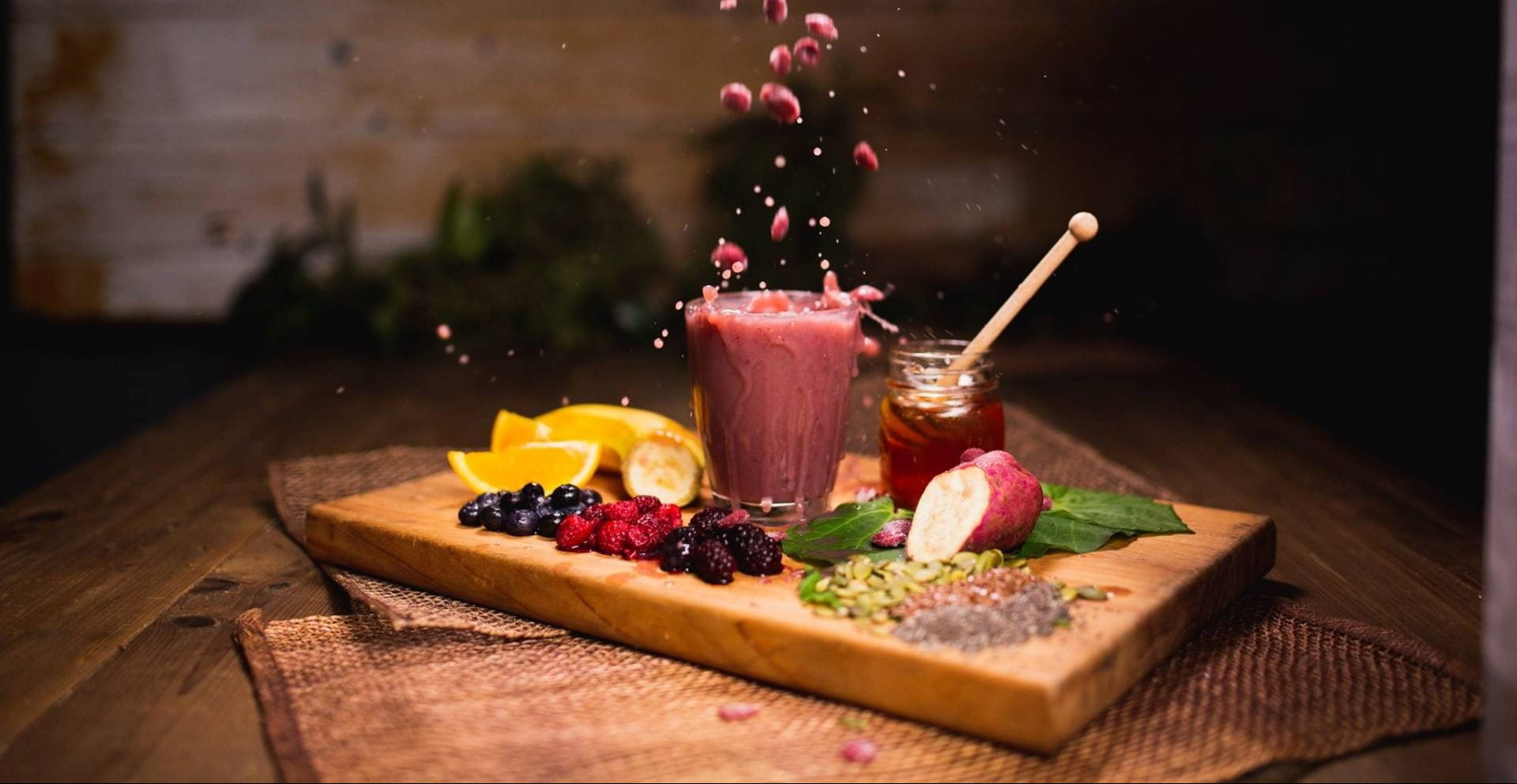 A pink smoothie is a glass standing on a wooden chopping board, alongside a jar of honey, seeds and freshly cut fruit